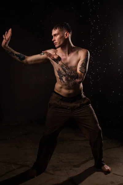 Tattooed man with water drops on his body, handsome healthy tattooed young man in the rain. Sexy fitness man. Naked torso with tattoos. Fashion male posing in studio on dark background.