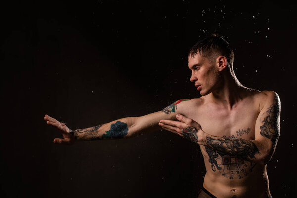 Tattooed man with water drops on his body, handsome healthy tattooed young man in the rain. Sexy fitness man. Naked torso with tattoos. Fashion male posing in studio on dark background.