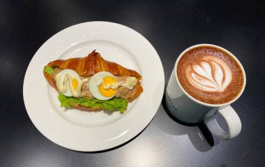 Çiçekli latte sanatıyla süslenmiş bir fincan taze kahve. Lezzetli mayonez ton balıklı tereyağlı ton balıklı sandviç, bir parça taze marul ve iki dilim haşlanmış yumurta.. 