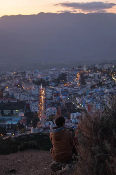 Genç adam gün batımında Fas 'ın Chefchaouen şehrinde sırtüstü oturup şehri ışıklarla izliyor..