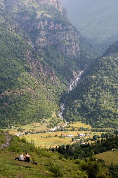 Panoramic view of the mountains with a river crossing them, and two girls resting, in the Mestia-Ushguli trek, in Georgia.