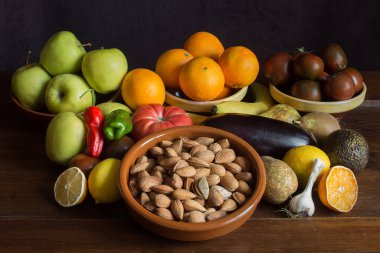Still life background with whole almonds, garlic, banana, oranges, peppers, avocado, tomato, lemon, pears, and apples on natural dark wood and clay bowls. Celebrations and healthy food.	