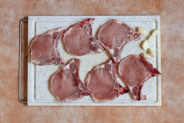 Вид сверху на свиные отбивные на разделочной доске на абстрактном художественном мраморном фоне с чесноком, готовым к приготовлению. Рецепты и потребление мяса.