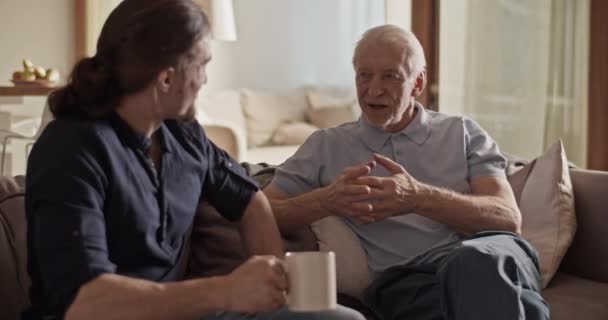 年事已高的父亲坐在沙发上和成年儿子聊天喝茶 — 图库视频影像