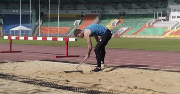 アスリートは長いジャンプトレーニング中にブラシでピットに砂を広げる — ストック動画