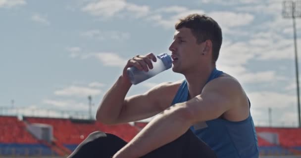 陸上競技場でトラックに座っている間に水をすすいで疲れた若いスポーツマン — ストック動画