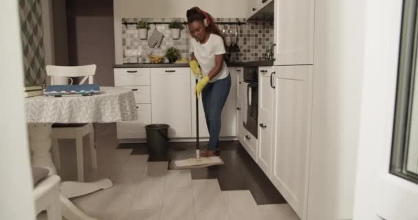 Afroamerikanerin Mit Kopfhörer Putzt Küchenboden Mit Wischmopp — Stockvideo