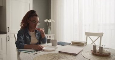 Black student biting toast and sipping coffee while writing down data