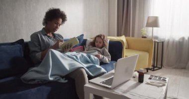 Black mother closing laptop and caressing ill daughter on sofa at home