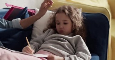 Black mom caressing focused kid drawing on paper on sofa at home