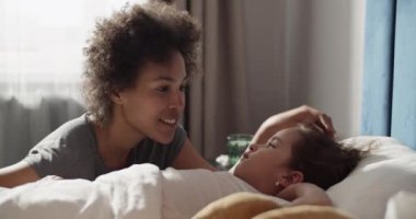Black mom smiling and caressing sick mixed race kid lying on bed