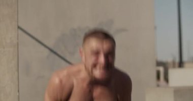 Üstsüz sporcu kükreyen ve antrenman sırasında kettlebell salıncakları yapan.