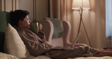 Female in bathrobe with sheet mask sipping tea from mug and watching film on netbook while chilling on bed during skin care routine in evening at home