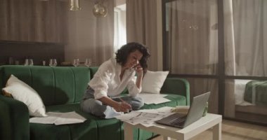 Adult woman in smart casual clothes checking drafts and data on laptop and speaking with customer via cellphone during remote work from home.