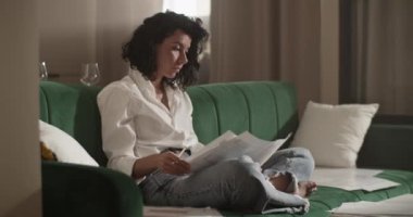 Barefoot adult woman in smart casual clothes writing and drawing on blueprints while sitting cross legged on couch and working on architectural project in living room at home.Full body footage