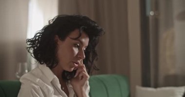 Female entrepreneur with dark hair discussing remote project with colleagues via cellphone while sitting on sofa in living room at home
