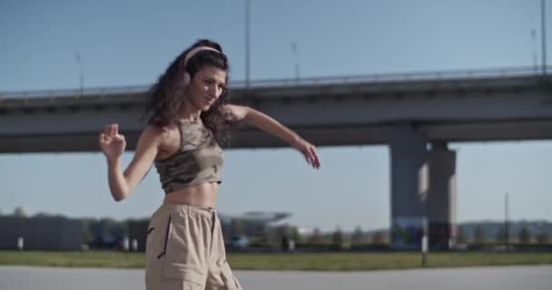 市内の晴れた朝に踊りながら ワイヤレスヘッドフォンで音楽を聴きながら肩を動かす若い女性ダンサーのハンドヘルドショット — ストック動画