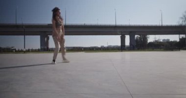 Çağdaş köprüye karşı beton zeminde müzik dinleyip dans eden enerjik genç bir kadının statik çekimi ve hafta sonu şehirde bulutsuz mavi gökyüzü.