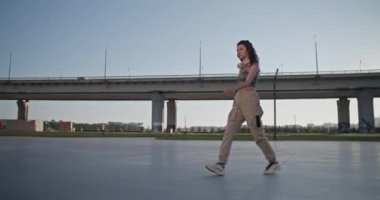 Bir şişe su ve kablosuz kulaklıkla, modern köprüye ve bulutsuz mavi gökyüzüne karşı yürüyen, sıradan giysiler giymiş genç bir kadının tüm vücudunun fotoğrafını çekiyorum.