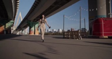 Güneşli bir sabahta şehir caddesinde modern köprünün altında bir şişe suyla gezen sıradan elbiseli bir kadının fotoğrafını çekiyorum.