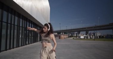 Koyu renk saçlı, kablosuz kulaklıkla müzik dinleyen ve bulutsuz bir sabah modern binanın yanında dans eden zarif genç bir kadının el kamerasıyla çekilmiş fotoğrafı.