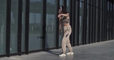 Şık giyinen, cam duvardaki yansımaya bakan ve şehirdeki prova sırasında kablosuz kulaklıkla müzik dinleyen kadının el kamerasıyla çekilmiş bir fotoğrafı.