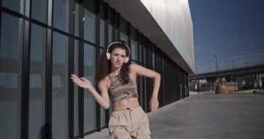 Gündüz kıyafetleri içinde genç bir kadının kollarını ve omuzlarını hareket ettirirken günümüzün modern binasının yakınında güneşli bir sabahta cam duvarla dans ederken çekilmiş bir fotoğrafı.