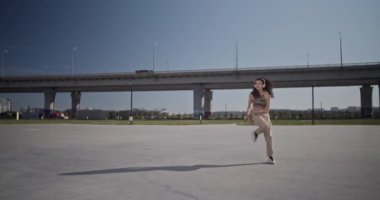 Şehrin güneşli sabahında modern hip hop dansı ve bulutsuz mavi gökyüzü performansı sergileyen, bütün vücutlu bir kadının el kamerası görüntüsü.