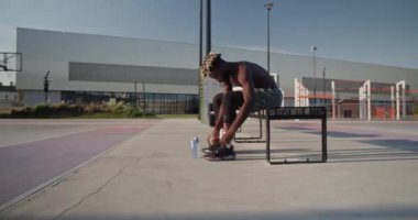Spor sahasında antrenmanlara hazırlanırken spor ayakkabılarını bağlayan, su ve topun yanındaki bankta oturan üstsüz Afrikalı Amerikalı basketbolcuya odaklan.