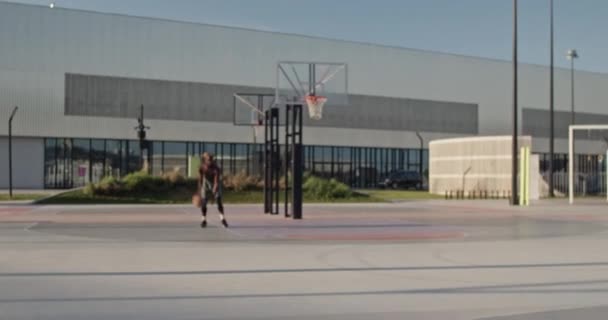 ズームビューで決定されたアフリカ系アメリカ人男性選手とともに裸の胴とブロンドのドリルボールとカメラに向かって実行しながら 日中のスポーツグラウンドでバスケットボール — ストック動画