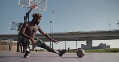 Düşük açılı, el kamerasıyla çekilmiş, üstsüz bir Afrikalı Amerikalı sporcunun, kentteki köprünün yakınındaki spor sahasında basketbol antrenmanına hazırlanırken topun yanına atlarken çekilmiş.