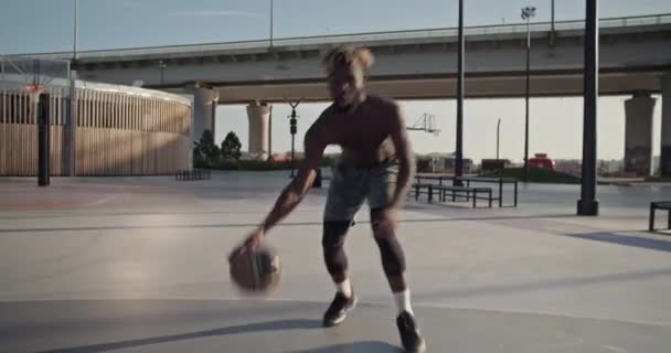 在城市阳光明媚的夏日 在桥边运动场上做篮球自由泳特技表演时 一名赤身裸体的非洲裔美国运动员正在运球的跟踪报道 — 图库视频影像