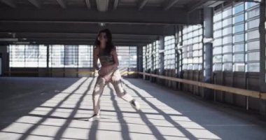 Güneş ışığında park yerinde dans ederken gündelik giysiler içinde müzik dinleyip kameraya bakan esmer bir kızı yakınlaştır.