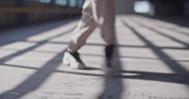 Tanımlanamayan bir dansçının sıradan pantolonu ve spor ayakkabılarıyla gündüz vakti kapalı otoparkta güneşli beton zeminde yürürken görüntüsü.