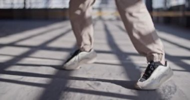 Tanımlanamayan bir dansçının sıradan pantolonu ve spor ayakkabılarıyla gündüz vakti kapalı otoparkta güneşli beton zeminde yürürken görüntüsü.
