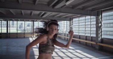 Gündelik giysiler ve kablosuz kulaklıklar takan, beton zeminde dans eden, müzik dinleyen, güneşli otoparkta dans eden ciddi genç bir kadının bakışını eğ.