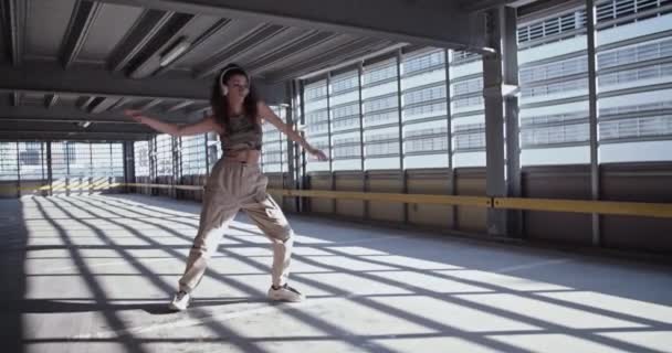 在空旷的室内停车场 穿着休闲装的年轻女子在阳光下听音乐和跳舞的手持镜头 — 图库视频影像