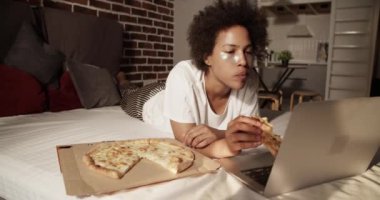 Afro-Amerikalı kadın evde film izlerken pizza yiyor.
