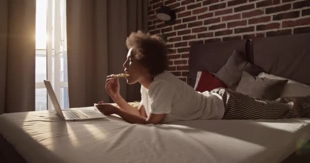 アフリカ系アメリカ人女性ですPajamaで巻き毛食べるピザとうなずきながら ベッドでリラックスしてネット上で映画を見て — ストック動画