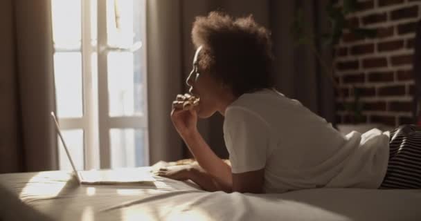 アフリカ系アメリカ人女性とともに巻き毛食べるピザと調整眼パッチながら横にベッドと見て映画上のラップトップ — ストック動画