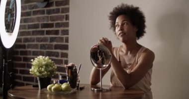 Kıvırcık saçlı, gözlerini kapayan ve yüzüne nemlendirici losyon sıkan siyah bir kadın masada oturmuş güzellik blogu için video kaydediyor.