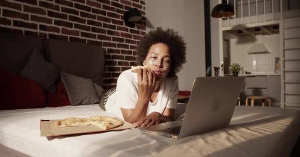 大人黒女性でベッドウェアでアイパッチチューニングおいしいピザと見ビデオ上のラップトップ — ストック動画