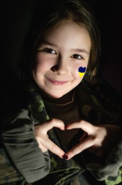 Yanağında Ukrayna bayrağı olan vatansever bir çocuk. Elleriyle yüreğini gösteriyor ve siyahlara saklanmış şekilde gülümsüyor. 