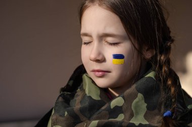 upset kid with painted ukrainian flag on cheek and camouflage jacket