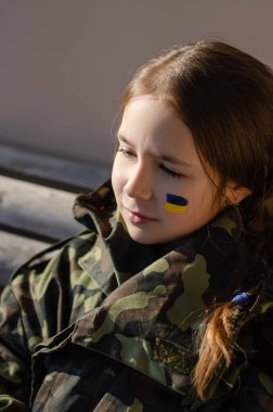 Yanağında Ukrayna bayrağı ve kamuflaj ceketi olan üzgün çocuk.