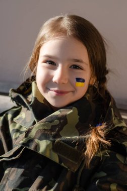 smiling kid with painted ukrainian flag on cheek and camouflage jacket