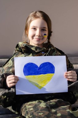 cheerful kid with painted ukrainian flag on cheek and camouflage jacket holding paper with heart and national flag colors