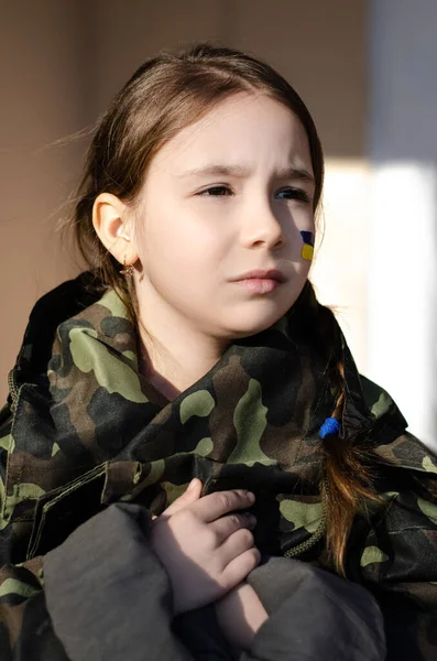 Niño triste con bandera pintada de Ucrania en la mejilla y camuflaje chaqueta - foto de stock
