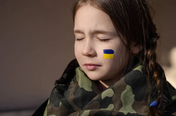 Niño molesto con bandera pintada de Ucrania en la mejilla y camuflaje chaqueta - foto de stock