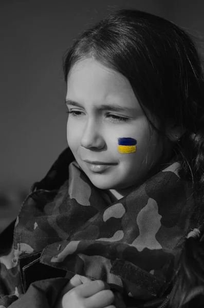 Monochrome photo of upset kid with painted ukrainian flag on cheek and camouflage jacket — Stock Photo